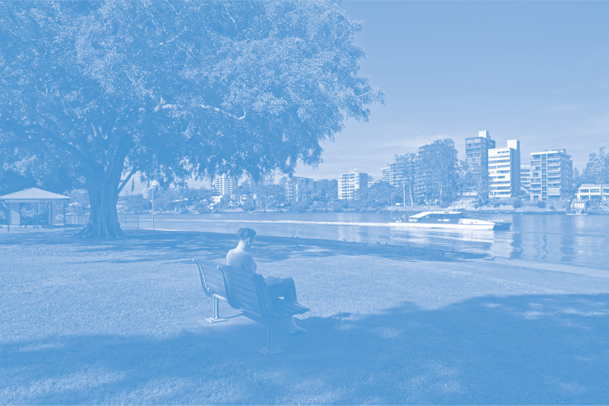 A person sits on a bench in a tranquil public park next to a river as a boat passes by.