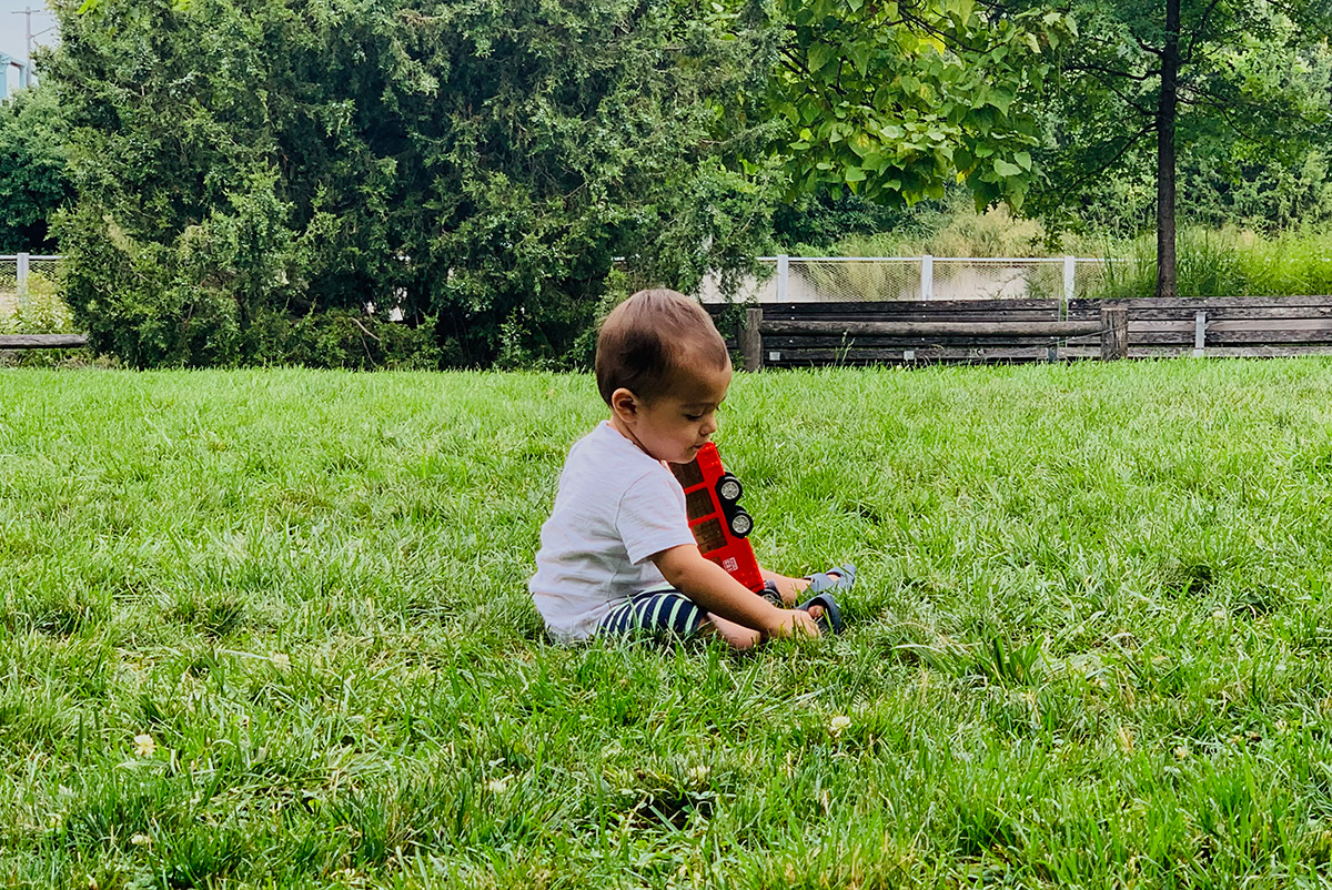 A small toddler sits in a lawn of green grass near some trees, playing with a toy firetruck.